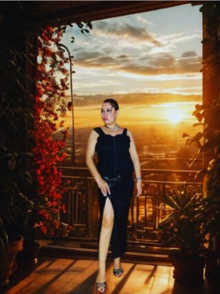 Curvy woman wearing a black corset slit dress on a balcony at sunset, perfect for elegant evening wear.