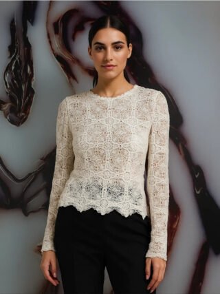 A woman wearing an off-white ivory long-sleeve geometric lace top with scalloped edges, paired with black trousers, standing against a marbled background.